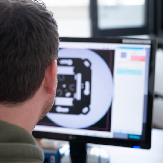 Symbolisch für den Komplettservice. Christian Friedenstab unscharf im Vordergrund beim Überwachung der Bauteilvermessung vor einer CAD-Ansicht.